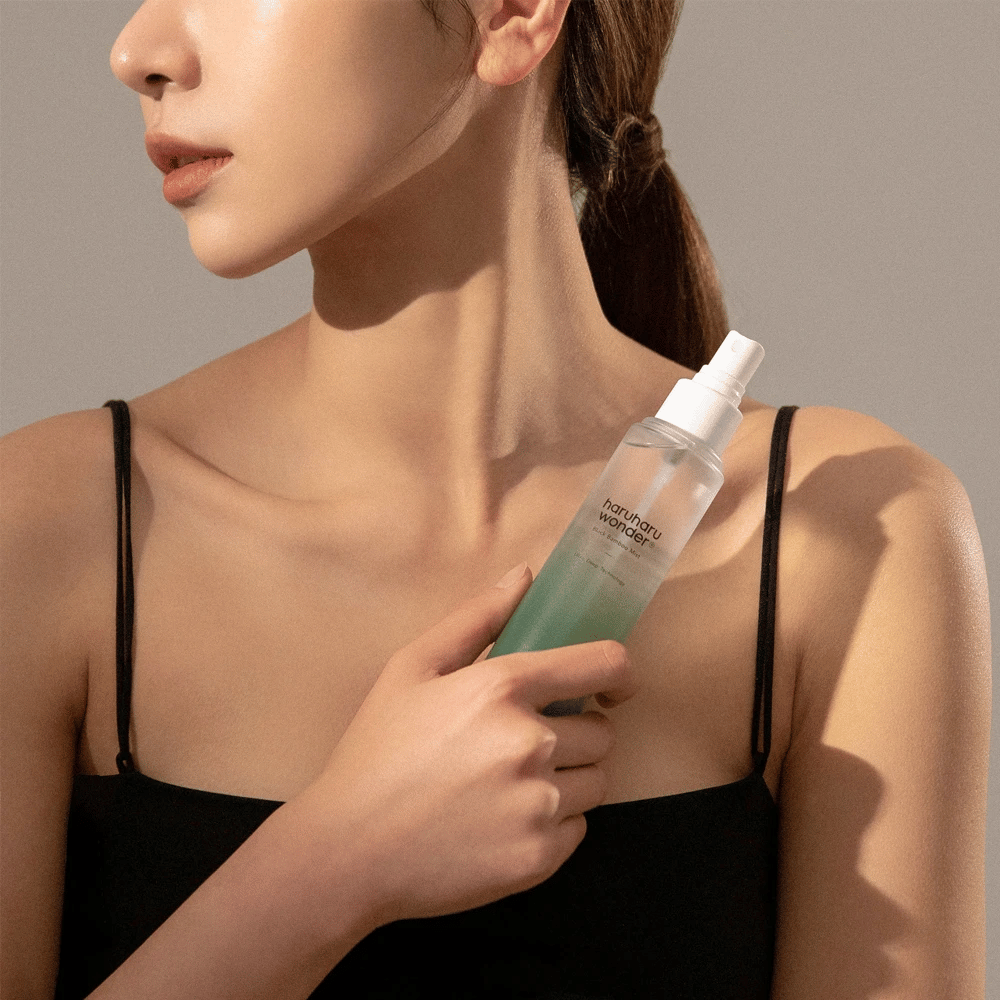 Slika HaruHaru Wonder Black Bamboo Mist, ki jo drži ženska. Izdelek je v prozorni razpršilni steklenički z zelenim odtenkom in belim razpršilnikom. Ženska ima sproščen videz, nosi črn top in se osredotoča na uporabo meglice za vlaženje in sijaj kože. Steklenička je del rutine nege kože.