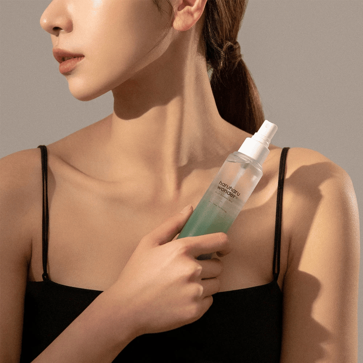 Slika HaruHaru Wonder Black Bamboo Mist, ki jo drži ženska. Izdelek je v prozorni razpršilni steklenički z zelenim odtenkom in belim razpršilnikom. Ženska ima sproščen videz, nosi črn top in se osredotoča na uporabo meglice za vlaženje in sijaj kože. Steklenička je del rutine nege kože.