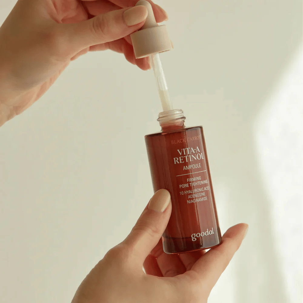 Roka drži stekleničko z Goodal Black Carrot Vita-A Retinol Firming Ampoule. Oznaka na steklenički prikazuje ime in lastnosti izdelka, vključno z "Učvrstitev in zategovanje por" ter sestavinami, kot so 10% hialuronska kislina, adenozin in niacinamid. Pipeta je v bež barvi.