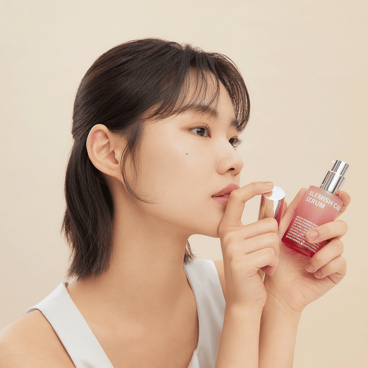 Ženska s temnimi lasmi drži stekleničko Blemish Care Serum v eni roki in s pokrovčkom v drugi. Ima nevtralen izraz na obrazu, ozadje pa je svetlo bež, kar daje mehki in minimalistični vtis.