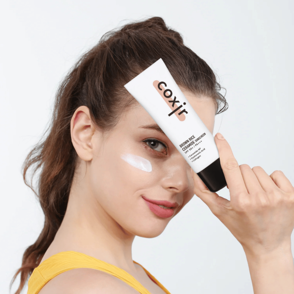Ženska z naneseno sončno kremo na licu drži tubo Coxir Brown Rice Ceramide Sunscreen, SPF 50+ PA+++, ob čelu in se rahlo smehlja.