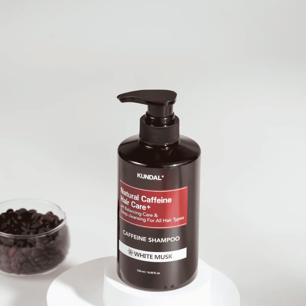 KUNDAL Natural Caffeine Hair Care+ Šampon White Musk v črni steklenici s pumpico, 500 ml, postavljen poleg sklede s kavnimi zrni.