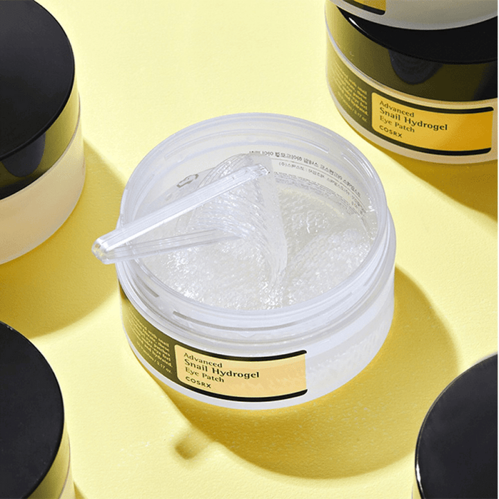 Odprta posoda COSRX Advanced Snail Hydrogel Eye Patch s prozornimi obliži za oči in majhno lopatico, obdana z več zaprtimi posodami na rumeni podlagi.