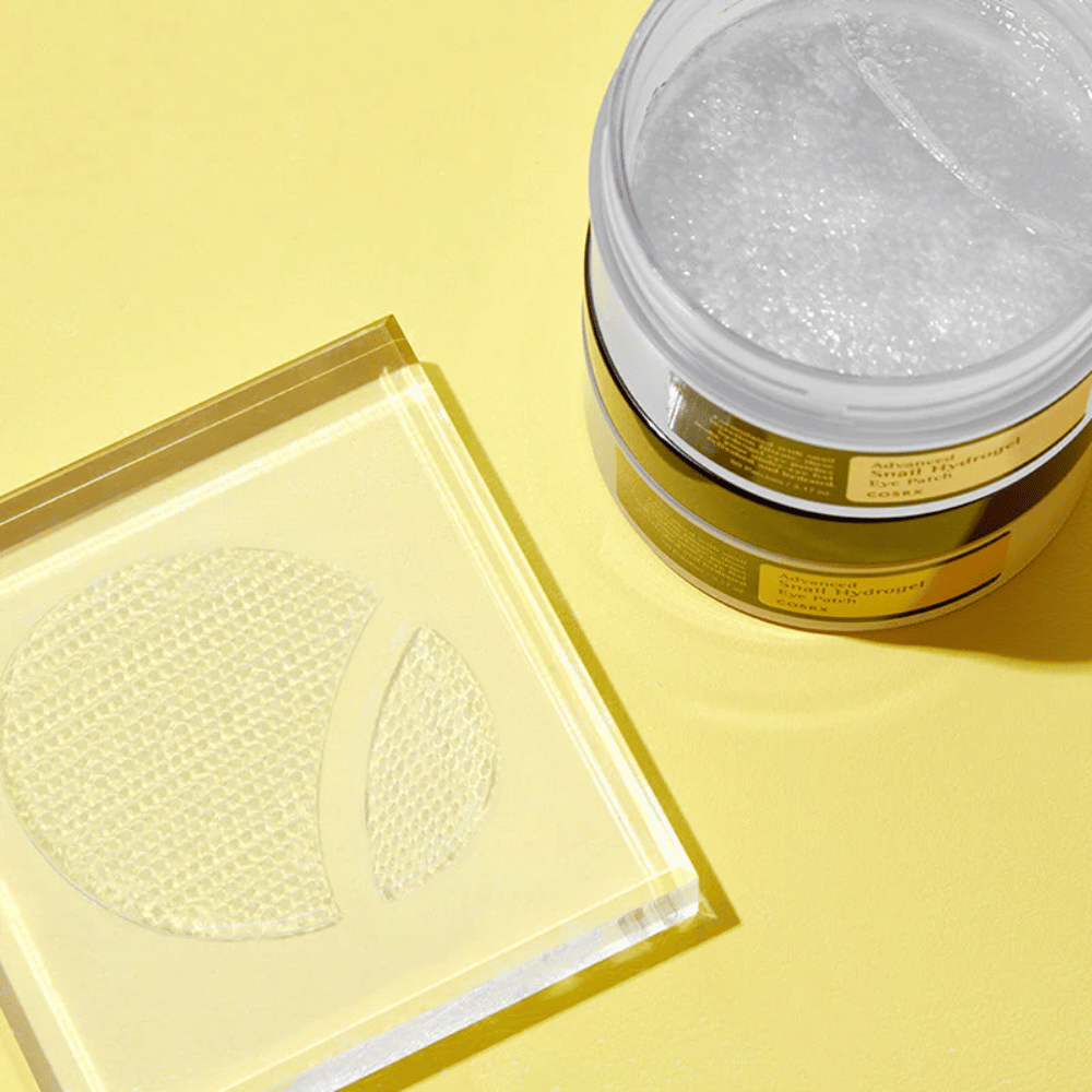 Odprta posoda COSRX Advanced Snail Hydrogel Eye Patch stoji na rumeni podlagi poleg prozorne podlage, kjer sta postavljena dva obliža za oči.