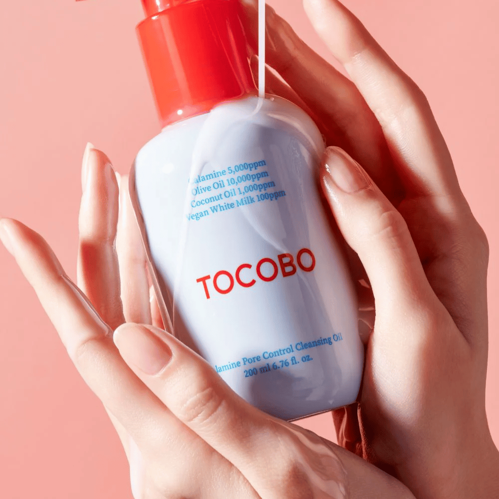 TOCOBO Calamine Pore Control Cleansing Oil drži v rokah proti roza ozadju, rdeča črpalka stekleničke in seznam sestavin sta jasno vidna.