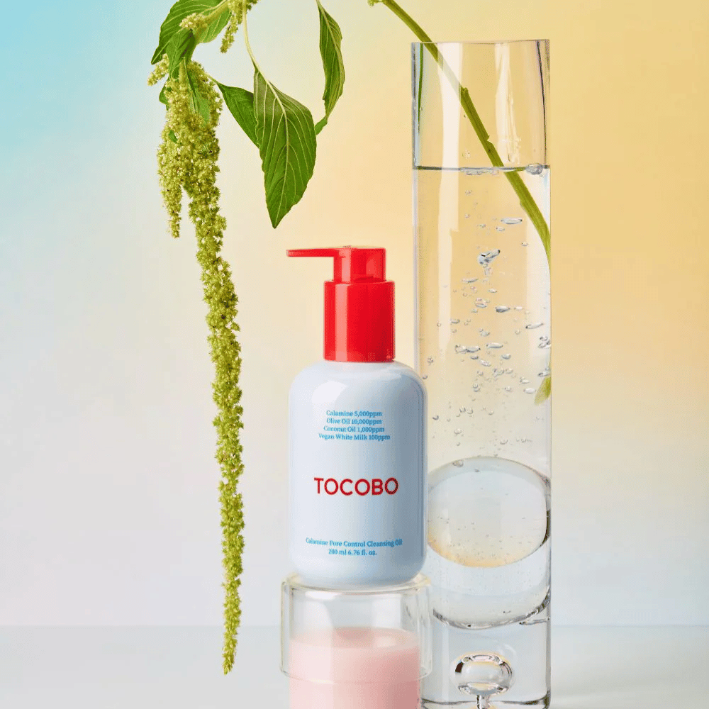 TOCOBO Calamine Pore Control Cleansing Oil z rdečo črpalko stoji poleg visokega steklenega valja, napolnjenega z vodo in zeleno rastlino, proti svetli pastelni barvni podlagi.