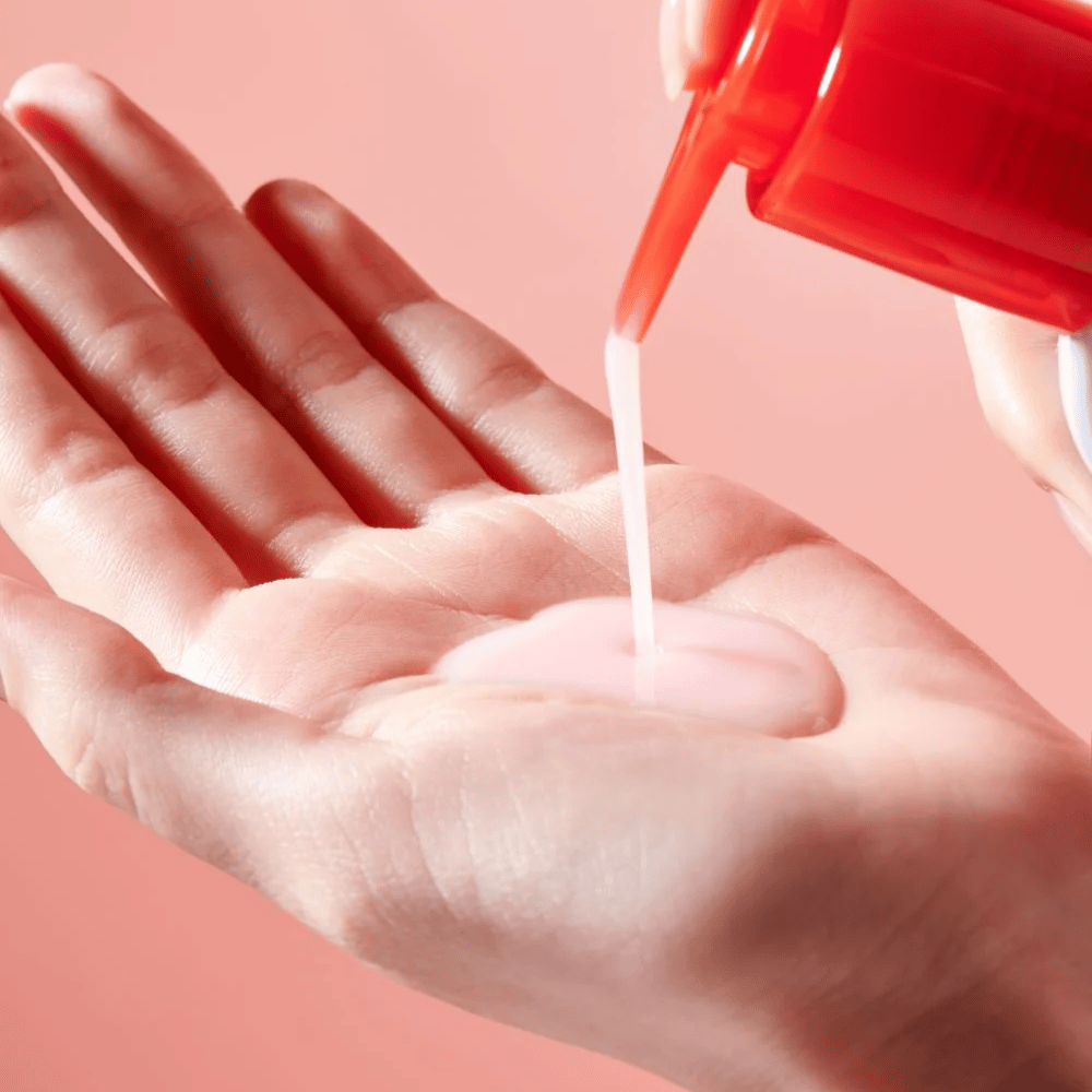 Svetlo rožnato čistilno olje se nanaša iz rdeče črpalke v odprto roko proti roza ozadju.