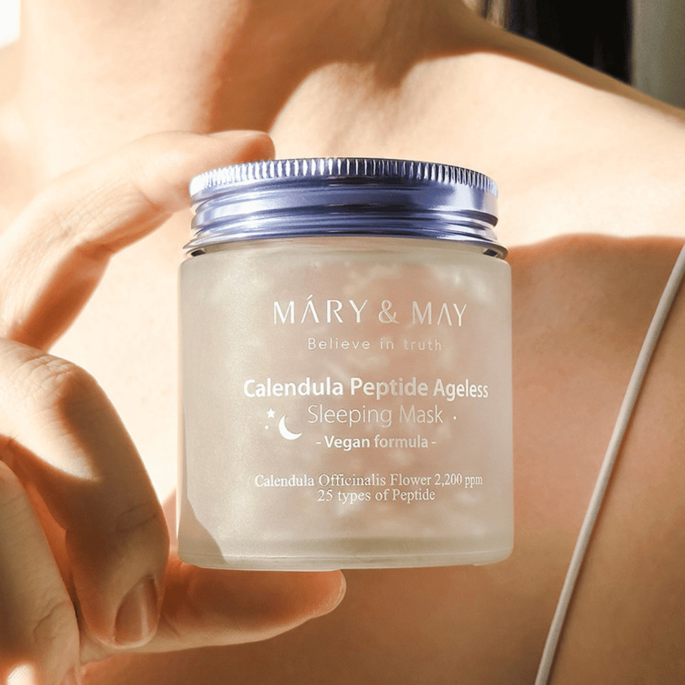 Ženska drži kozarec z MARY & MAY Calendula Peptide Ageless Sleeping Mask. Kozarec je prozoren z vijoličnim pokrovčkom in napis "Calendula Peptide Ageless Sleeping Mask" je jasno viden. Izdelek je veganska nočna maska z ognjičem in peptidi, namenjena dajanju mladostnega sijaja koži ponoči.