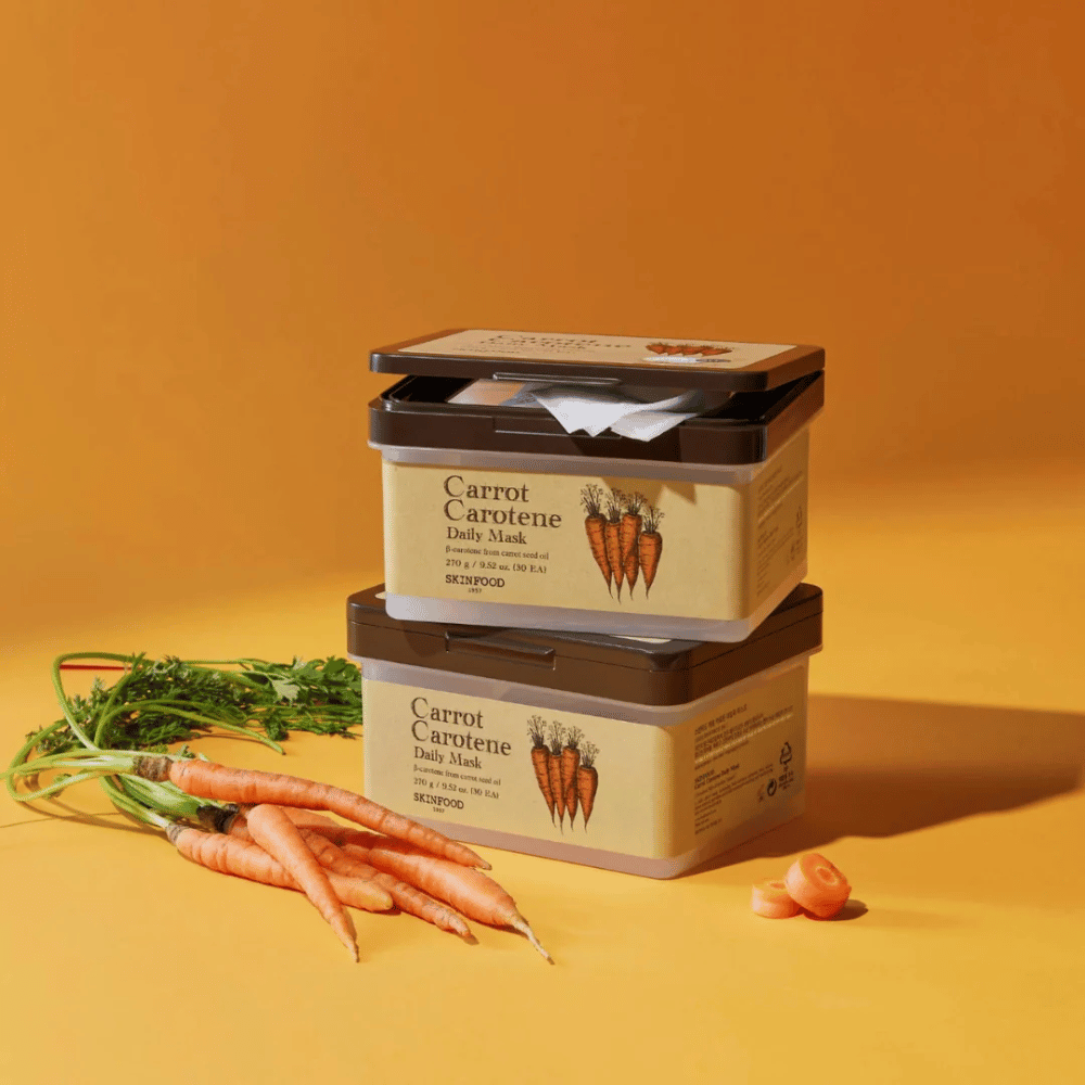 Slika prikazuje tri naložene pakete "Carrot Carotene Daily Mask" iz SKINFOOD, postavljene na oranžno ozadje skupaj s svežim korenjem. Vsaka škatla vsebuje obrazne maske, obogatene z β-karotenom iz olja korenčkovih semen, zasnovane za vsakodnevno uporabo za nego in negovanje kože.