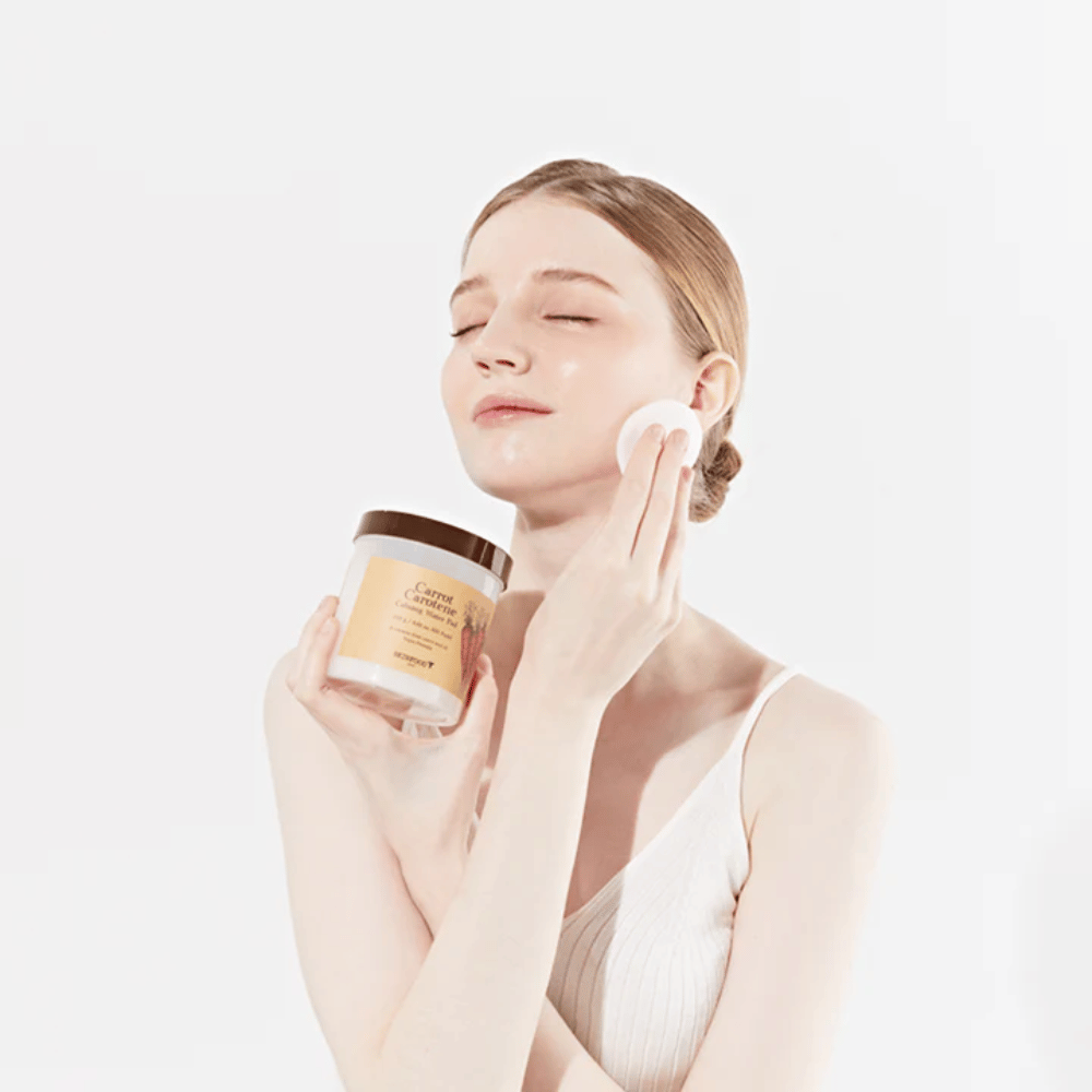 Ženska s spetimi lasmi nanese izdelek za nego kože na svoj obraz z bombažno blazinico. Drži veliko posodo z oznako "Carrot Carotene Calming Water Pad". Njene zaprte oči in miren izraz izražajo občutek sprostitve in uživanja v rutini nege kože.