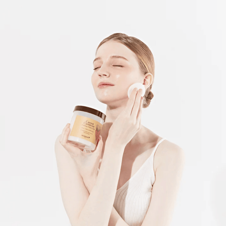 Ženska s spetimi lasmi nanese izdelek za nego kože na svoj obraz z bombažno blazinico. Drži veliko posodo z oznako "Carrot Carotene Calming Water Pad". Njene zaprte oči in miren izraz izražajo občutek sprostitve in uživanja v rutini nege kože.