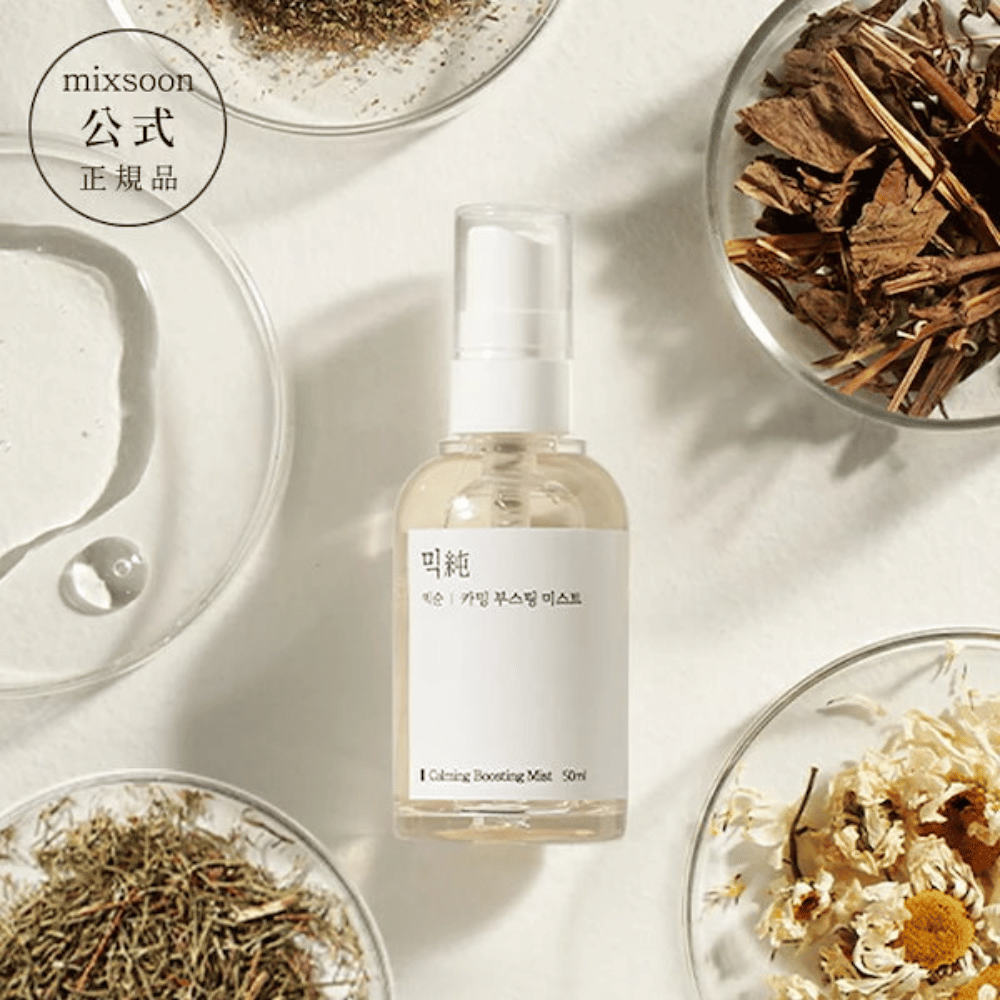 Prozorna razpršilna steklenička z etiketo 'Calming Boosting Mist' od Mixsoon, obdana s posušenimi zelišči in cvetovi na belih skledah proti bež ozadju, kar poudarja naravne sestavine izdelkov.