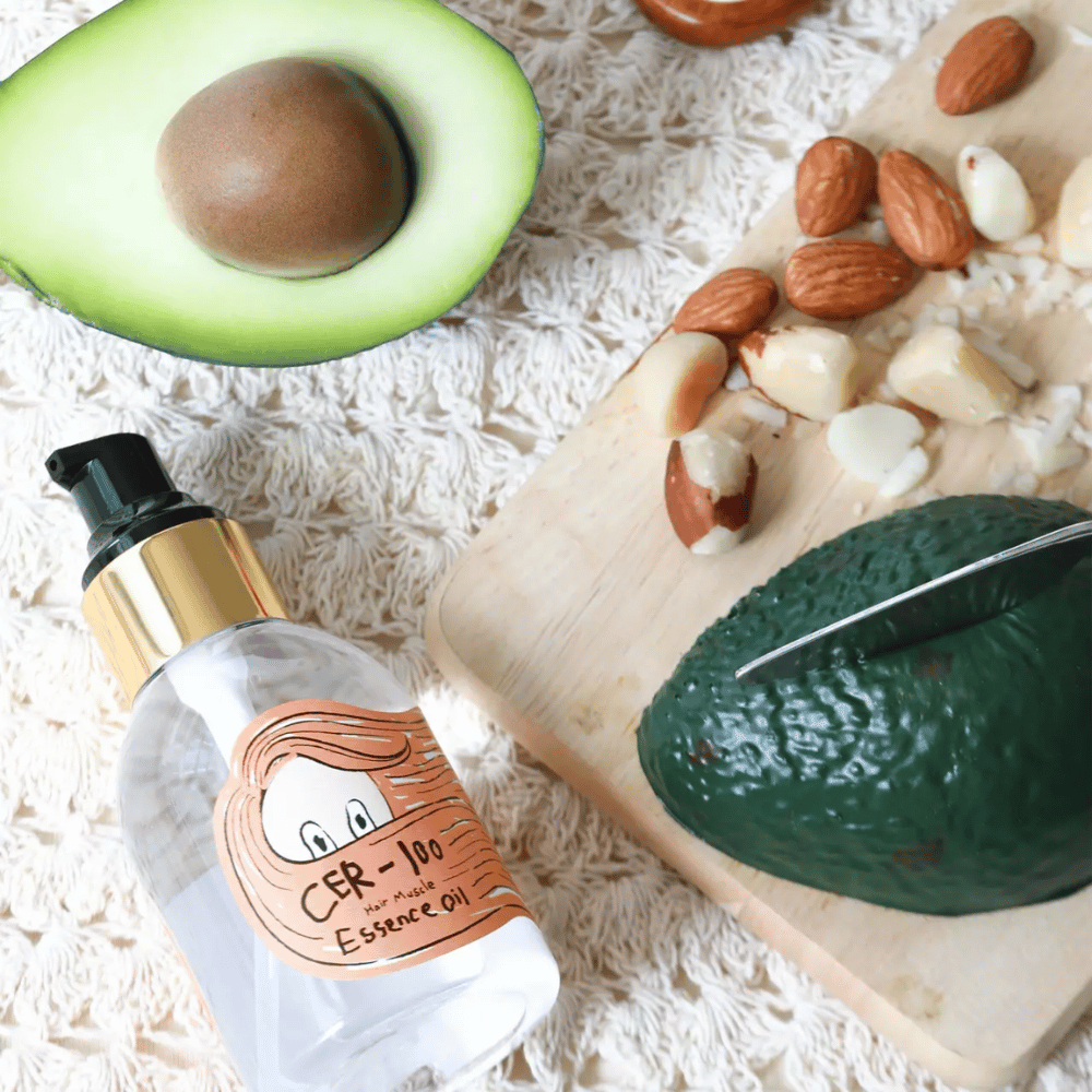 Elizavecca CER-100 Hair Muscle Essence Oil sestavine – bližnji posnetek z avokadom in rastlinskimi olji, ki prikazuje hranljivo olje za lase, ki pomaga mehčati in negovati lase.