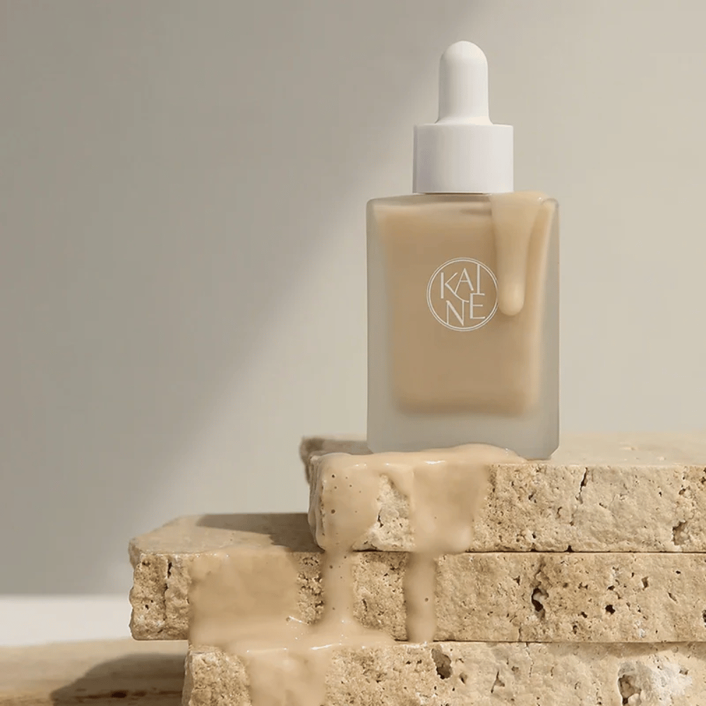 KANE Chaga Collagen Charging Serum v zmrznjeni stekleni steklenički s pipeto. Kremasta tekstura teče po naravnih kamnih v mehki in minimalistični svetlobi.