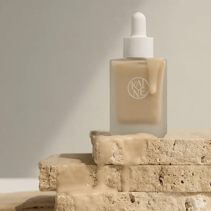 KANE Chaga Collagen Charging Serum v zmrznjeni stekleni steklenički s pipeto. Kremasta tekstura teče po naravnih kamnih v mehki in minimalistični svetlobi.