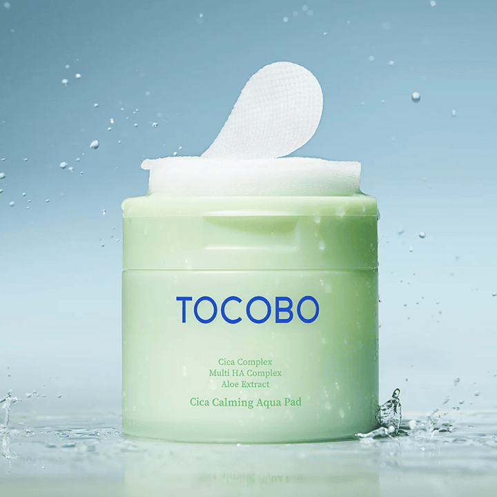 Odprt lonček z TOCOBO Cica Calming Aqua Pad, kjer iz embalaže štrli ena tonik za obraz blazinica. Kapljice vode okoli izdelka poudarjajo občutek intenzivne hidracije in svežine.