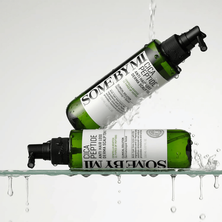 Dve steklenici SOME BY MI Cica Peptide Anti Hair Loss Derma Scalp Tonic ležita v in nad vodo s kapljicami, ki škropijo, zelene steklenice s črnim razpršilnim nastavkom in belo etiketo.