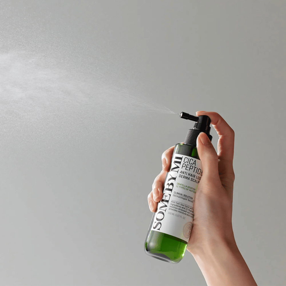 Roka, ki razprši SOME BY MI Cica Peptide Anti Hair Loss Derma Scalp Tonic iz zelene steklenice s črnim nastavkom in belo etiketo proti sivi podlagi.