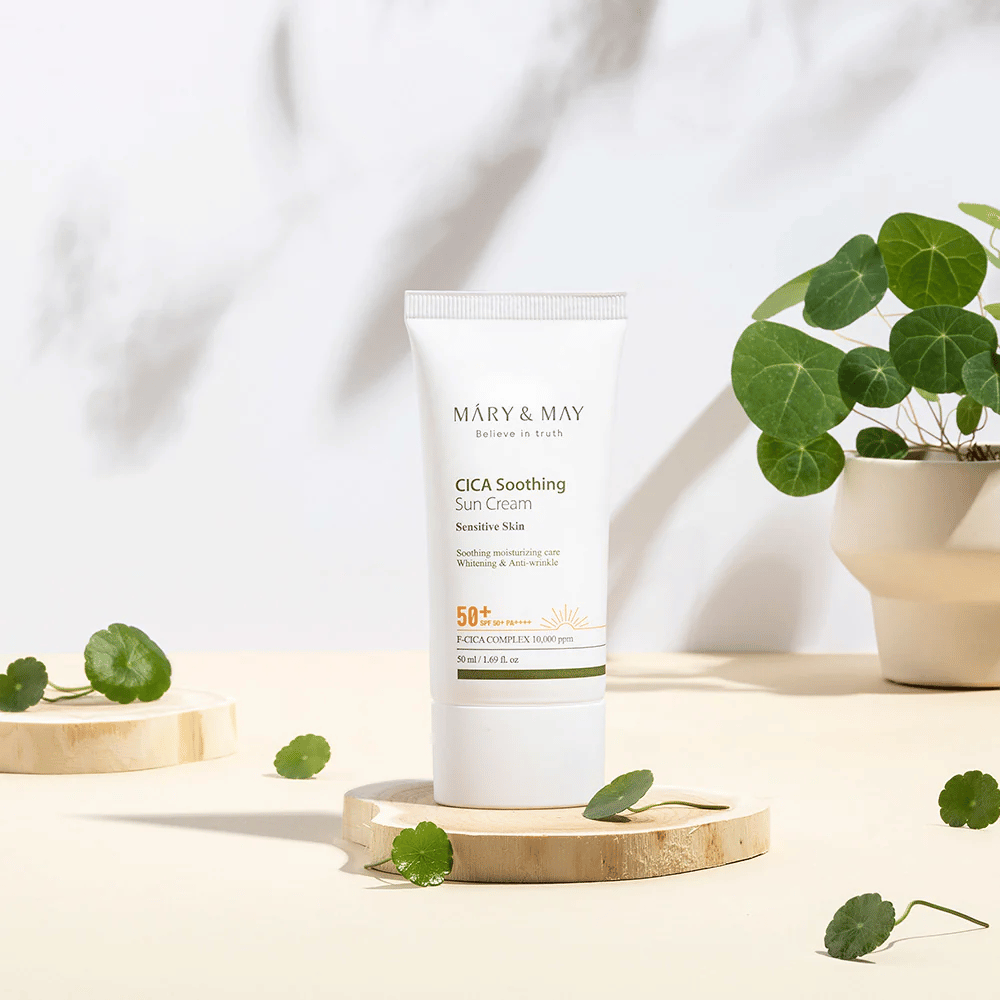 Mary&May CICA Soothing Sun Cream postavljen na lesen podstavek z zelenimi listi centelle okoli njega. Slika poudarja pomirjujoče sestavine za kožo in naravni navdih izdelka.