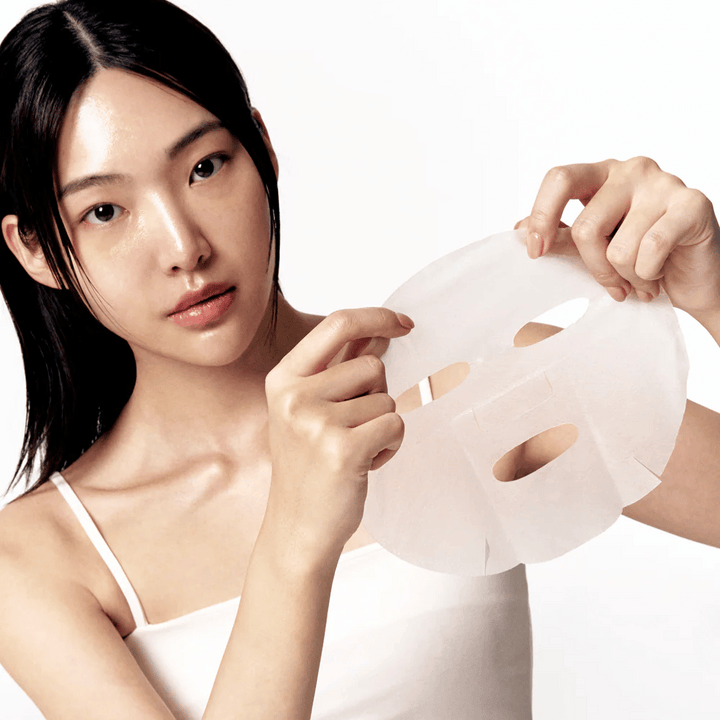 Ženska s čisto in navlaženo kožo drži belo masko iz listov pred seboj, pripravljena, da jo nanese na obraz.