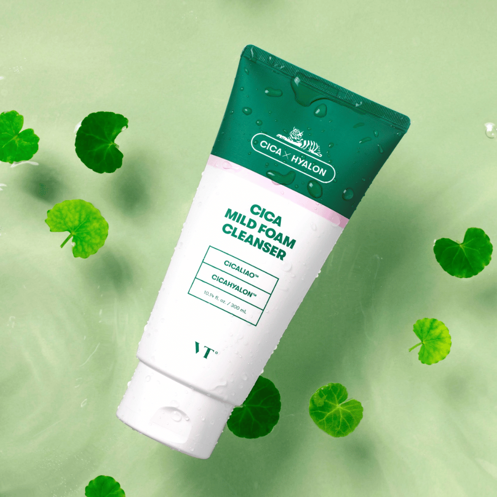 VT Cica Mild Foam Cleanser leži na svetlo zeleni podlagi z vodnimi kapljicami na embalaži in obkrožen z zelenimi listi centella asiatica, kar poudarja pomirjujoče lastnosti izdelka.