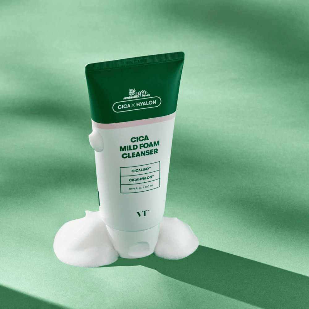 Tuba z VT Cica Mild Foam Cleanser stoji na zeleni podlagi z peno okoli dna. Izdelek ima zeleno in belo obliko ter se trži z CICALIAO™ in CICAHYALON™.