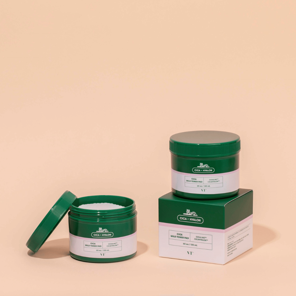 Dve zeleni posodi z VT Cica Mild Toner Pad, ena odprta, ki prikazuje bele blazinice za toniranje, in ena zaprta, postavljena na embalažo izdelka, na bež podlagi.