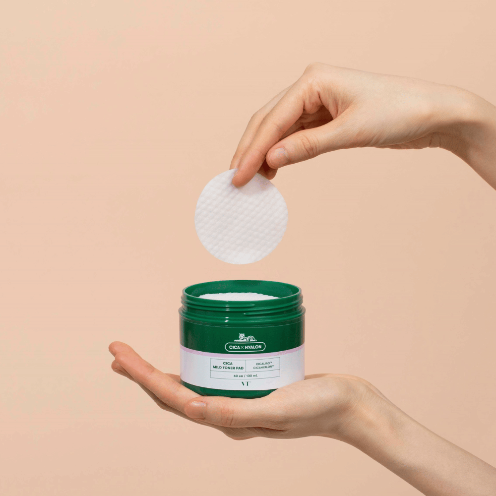 Roka, ki drži zeleno posodo z VT Cica Mild Toner Pad in dviguje okroglo, belo blazinico za toniranje nad posodo na bež podlagi.