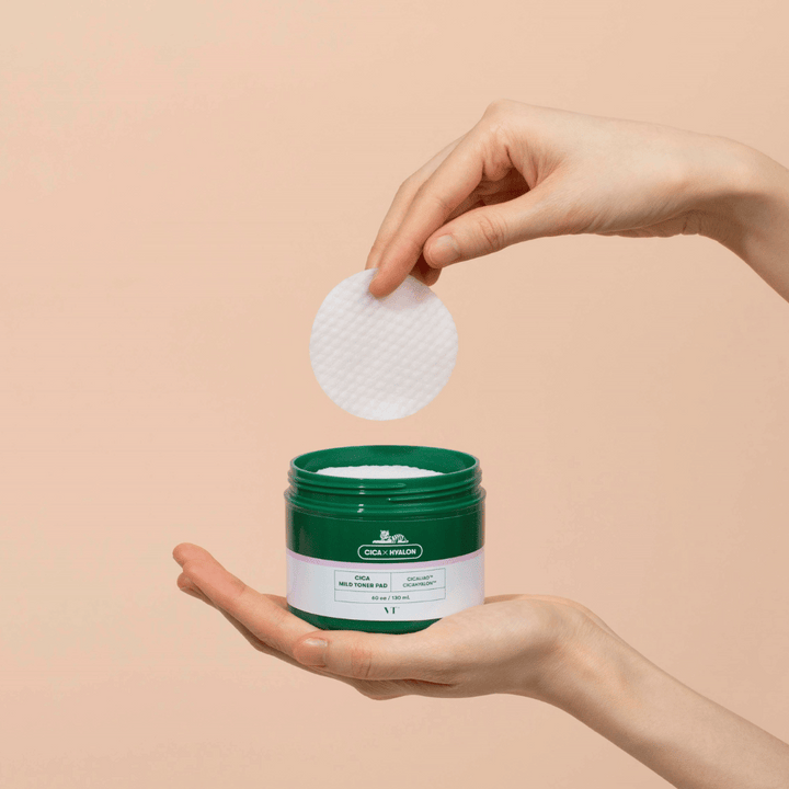 Roka, ki drži zeleno posodo z VT Cica Mild Toner Pad in dviguje okroglo, belo blazinico za toniranje nad posodo na bež podlagi.