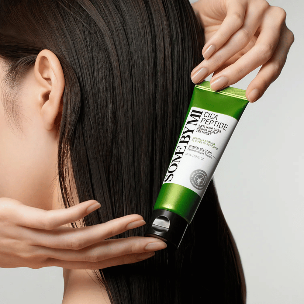 Ženska nanese SOME BY MI Cica Peptide Anti Hair Loss Derma Scalp Treatment v dolge, temne lase z zeleno in belo tubo.