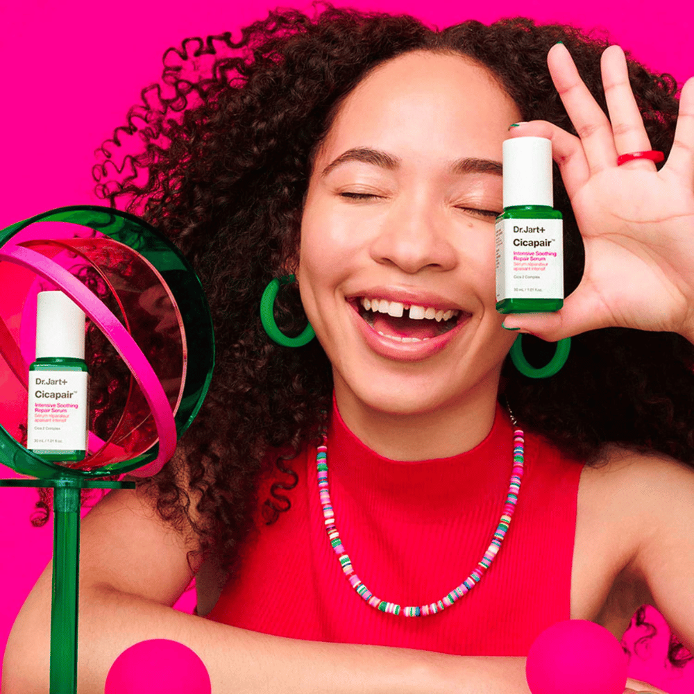 Ženska se široko smeji, medtem ko drži Dr.Jart+ Cicapair Intensive Soothing Repair Serum pred svojim obrazom. Nosi zelene uhane in rdečo ogrlico. Ozadje je svetlo roza in okrašeno z zelenimi in roza krogi.