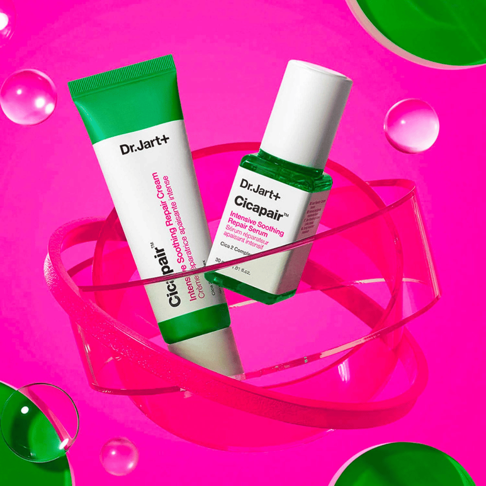 Dr.Jart+ Cicapair Intensive Soothing Repair Cream in Serum v zelenih embalažah, ki stojijo na roza krožni osnovi. Izdelki so zasnovani za pomiritev in obnovo razdražene kože.