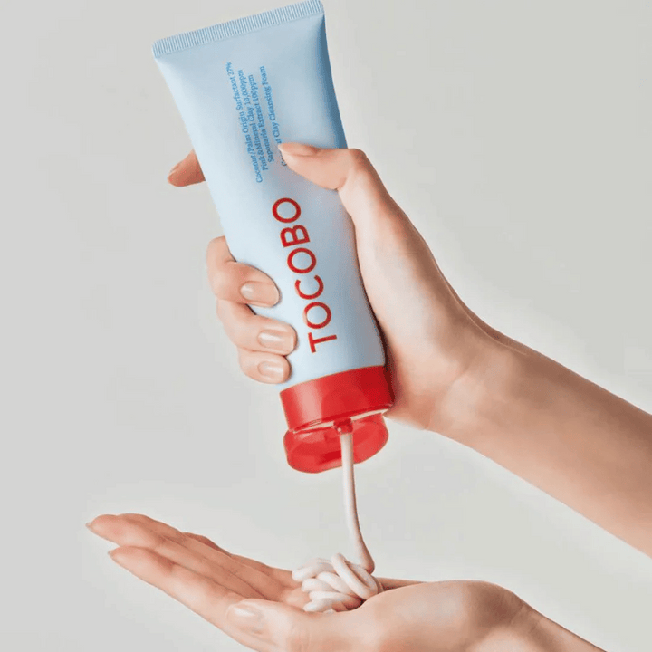Oseba drži tubo TOCOBO Coconut Clay Cleansing Foam. Tuba je svetlo modra z rdečim pokrovčkom in napisom "TOCOBO" na sprednji strani. Izdelek se iztisne iz tube in oblikuje kot šampon v roki. Ozadje je nevtralno in usmerja pozornost na izdelek.