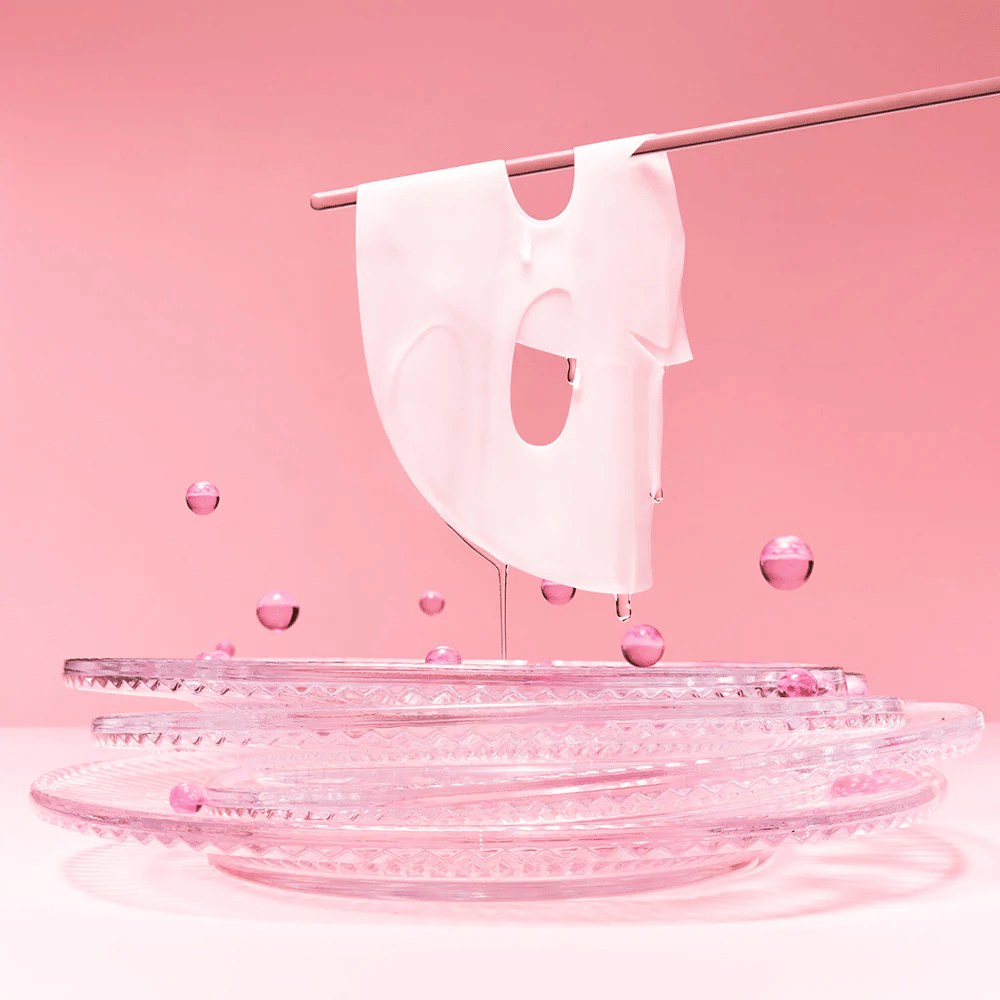 Hidrogelna maska visi na palici, kaplja s serumom. Ozadje je roza, ogledalo na dnu pa odseva roza mehurčke, ki obdajajo masko.