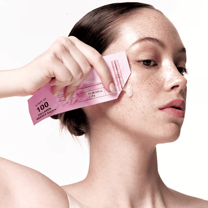 Ženska nanaša korak 1 Collagen Reedle Shot 100 z roza enkratno embalažo na lice. Jasni gel je viden na koži, kar označuje prvi korak dvostopenjske nege kože.