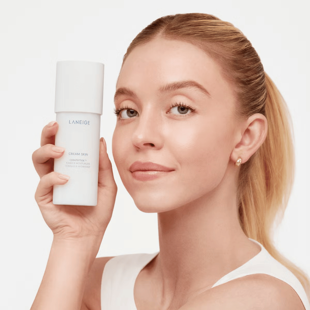 Oseba drži Laneige Cream Skin Cerapeptide Refiner v beli steklenički. Oseba se smehlja in prikazuje izdelek, ki se trži kot vlažilni toner in izdelek za nego kože.