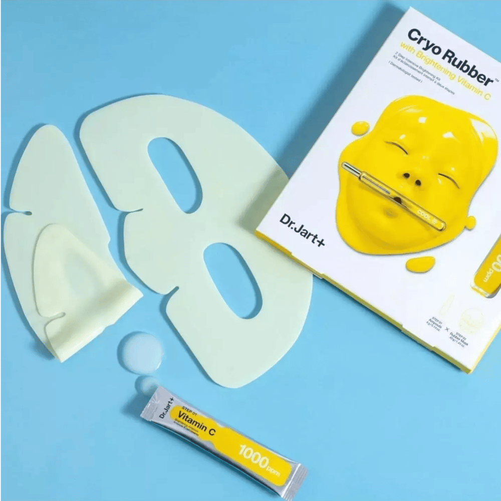Slika lepotnega izdelka Dr.Jart+'s 'Cryo Rubber™ z osvetljevalnim vitaminom C'. Na modrem ozadju je bela gumijasta obrazna maska, majhna tuba z oznako 'Vitamin C 1000ppm' in škatla z imenom izdelka ter sliko rumene maske.