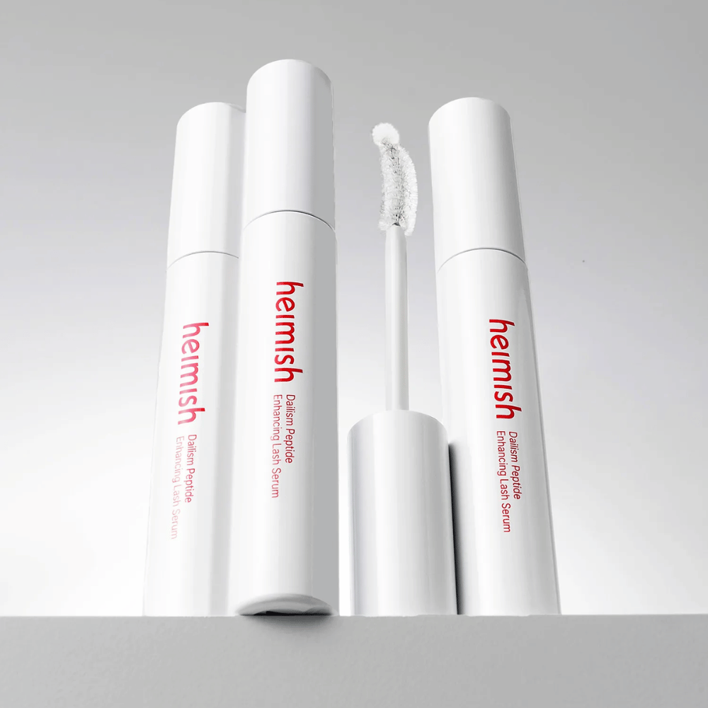 Več embalaž izdelka Heimish Dailism Peptide Enhancing Lash Serum je prikazanih skupaj z ukrivljeno aplikatorsko krtačko. Slika poudarja eleganten dizajn izdelka in natančno krtačko za enostaven nanos.