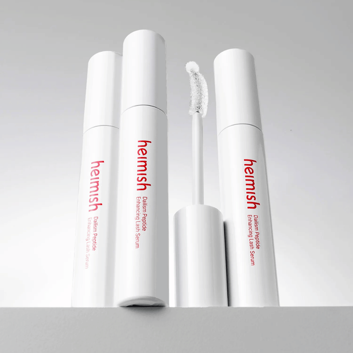 Več embalaž izdelka Heimish Dailism Peptide Enhancing Lash Serum je prikazanih skupaj z ukrivljeno aplikatorsko krtačko. Slika poudarja eleganten dizajn izdelka in natančno krtačko za enostaven nanos.