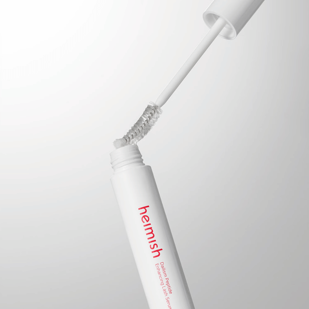Bližnji posnetek aplikatorja za Heimish Dailism Peptide Enhancing Lash Serum. Krtačka se izvleče iz bele posodice in prikazuje prozorno formulo seruma, ki se nanaša na trepalnice.