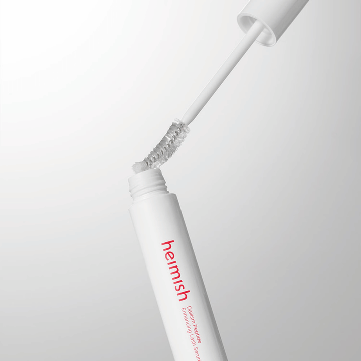 Bližnji posnetek aplikatorja za Heimish Dailism Peptide Enhancing Lash Serum. Krtačka se izvleče iz bele posodice in prikazuje prozorno formulo seruma, ki se nanaša na trepalnice.