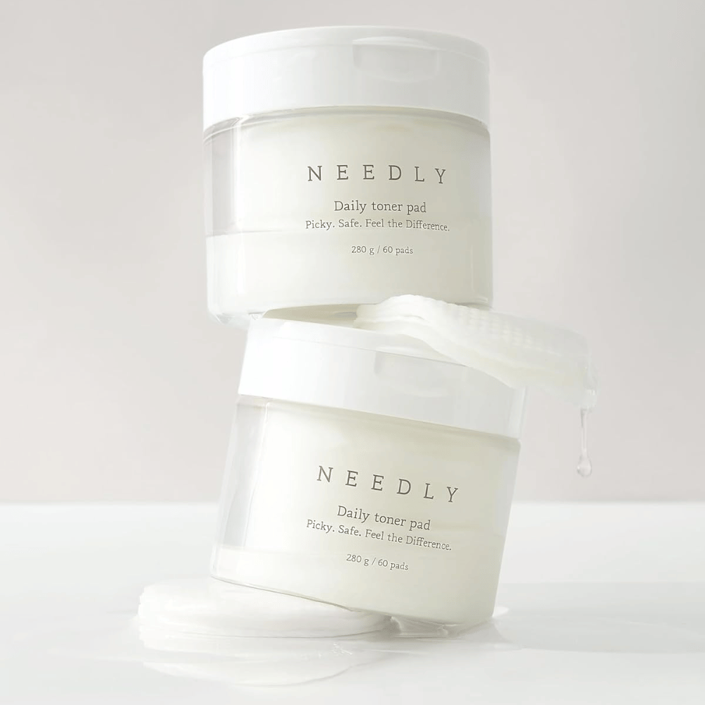 Dve posodi z Needly Daily Toner Pad, zloženi ena na drugo. Posode so bele z minimalističnim tekstom in vsebujejo po 60 tonerpads vsaka. Ena tonerpad delno leži zunaj zgornje posode s tekočino, ki kaplja.