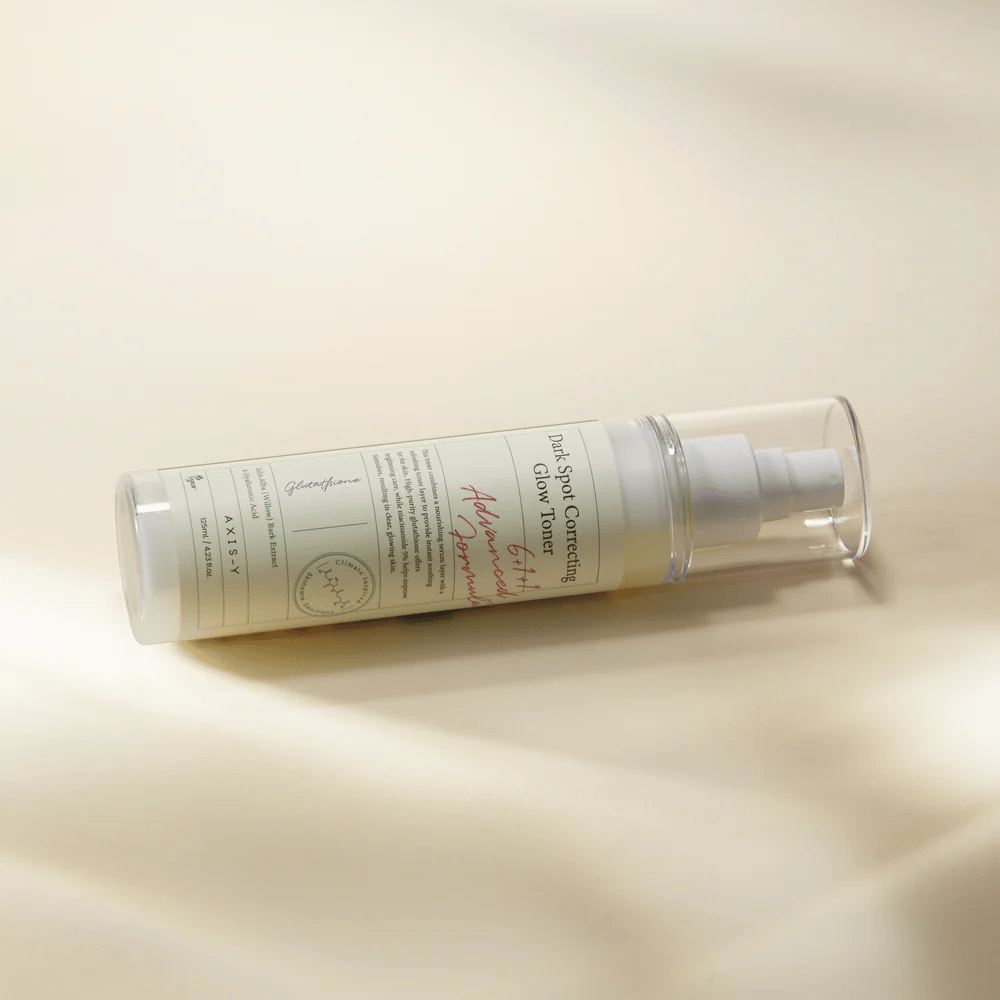Ležeča steklenica "AXIS-Y Dark Spot Correcting Glow Toner" postavljena na svetlo površino. Steklenica je prozorna z belo etiketo in ima belo razpršilno črpalko. Etiketa prikazuje informacije o svetlečih in vlažilnih lastnostih izdelka ter vsebuje ključne sestavine, kot sta glutathion in niacinamid.