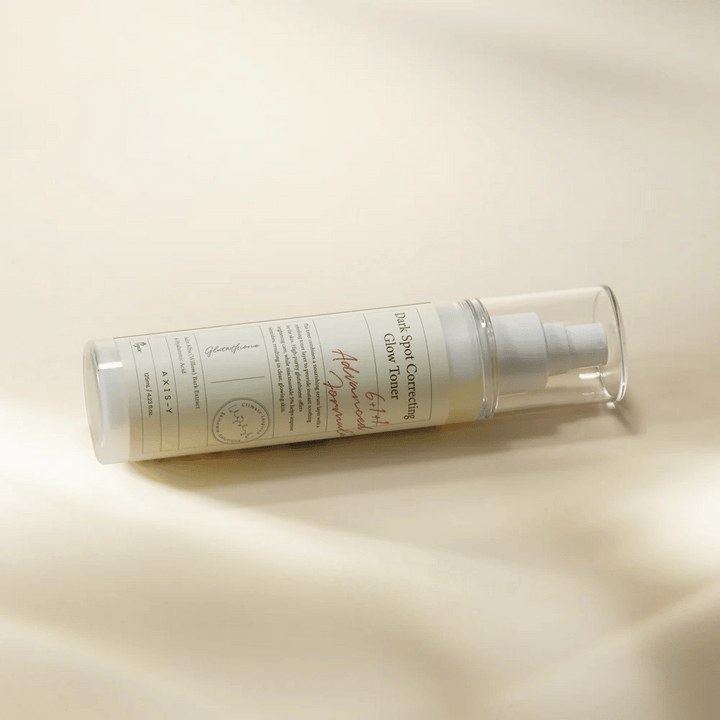 Ležeča steklenica "AXIS-Y Dark Spot Correcting Glow Toner" postavljena na svetlo površino. Steklenica je prozorna z belo etiketo in ima belo razpršilno črpalko. Etiketa prikazuje informacije o svetlečih in vlažilnih lastnostih izdelka ter vsebuje ključne sestavine, kot sta glutathion in niacinamid.