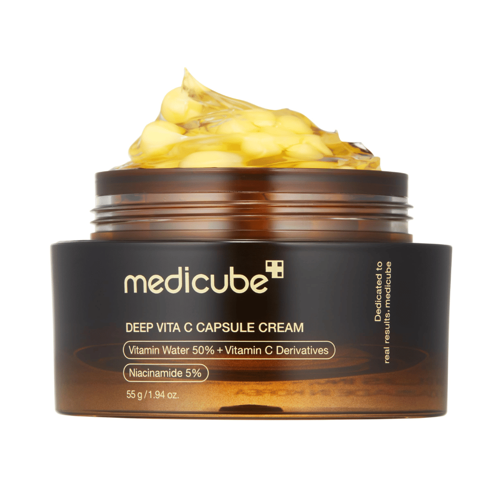 Bližnji posnetek "Medicube Deep Vita C Capsule Cream," ki prikazuje konsistenco kreme in kapsule z vitaminom C. Embalaža je temno rjava z zlatim besedilom in opisuje vsebino vitaminske vode (50 %) in niacinamida (5 %), kar daje vizualno predstavitev vlažilnih in izboljševalnih lastnosti izdelka.