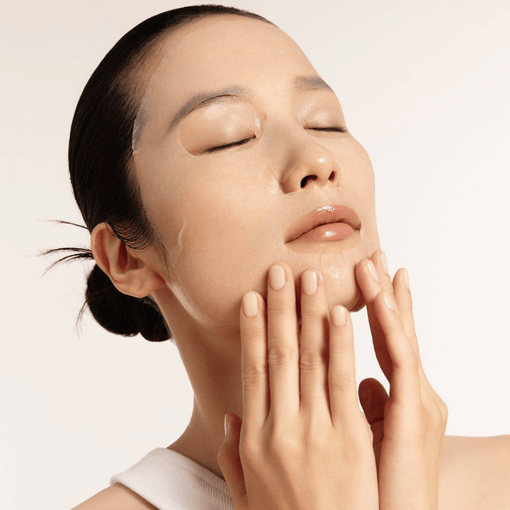 Model z navlaženo in sijočo kožo nanaša sheet masko, poudarek na sijaju, enakomerna polt in sijaj.