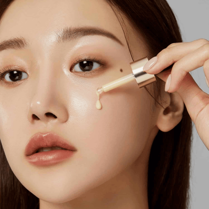 Bližnji posnetek ženske, ki nanaša Medicube Deep Vita C Ampoule z zlato pipeto. Kapljica seruma je na poti, da doseže njeno kožo, ki ima naravni sijaj. Popolno za svetlejši in enakomernejši ten kože.
