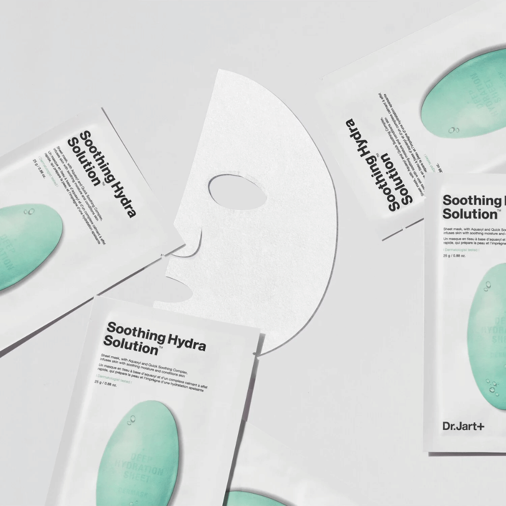 Več pakiranj obraznih mask "Soothing Hydra Solution" od Dr.Jart+ je razporejenih na svetli površini. Razvita maska je postavljena na sredino, kar prikazuje njeno belo in perforirano strukturo.