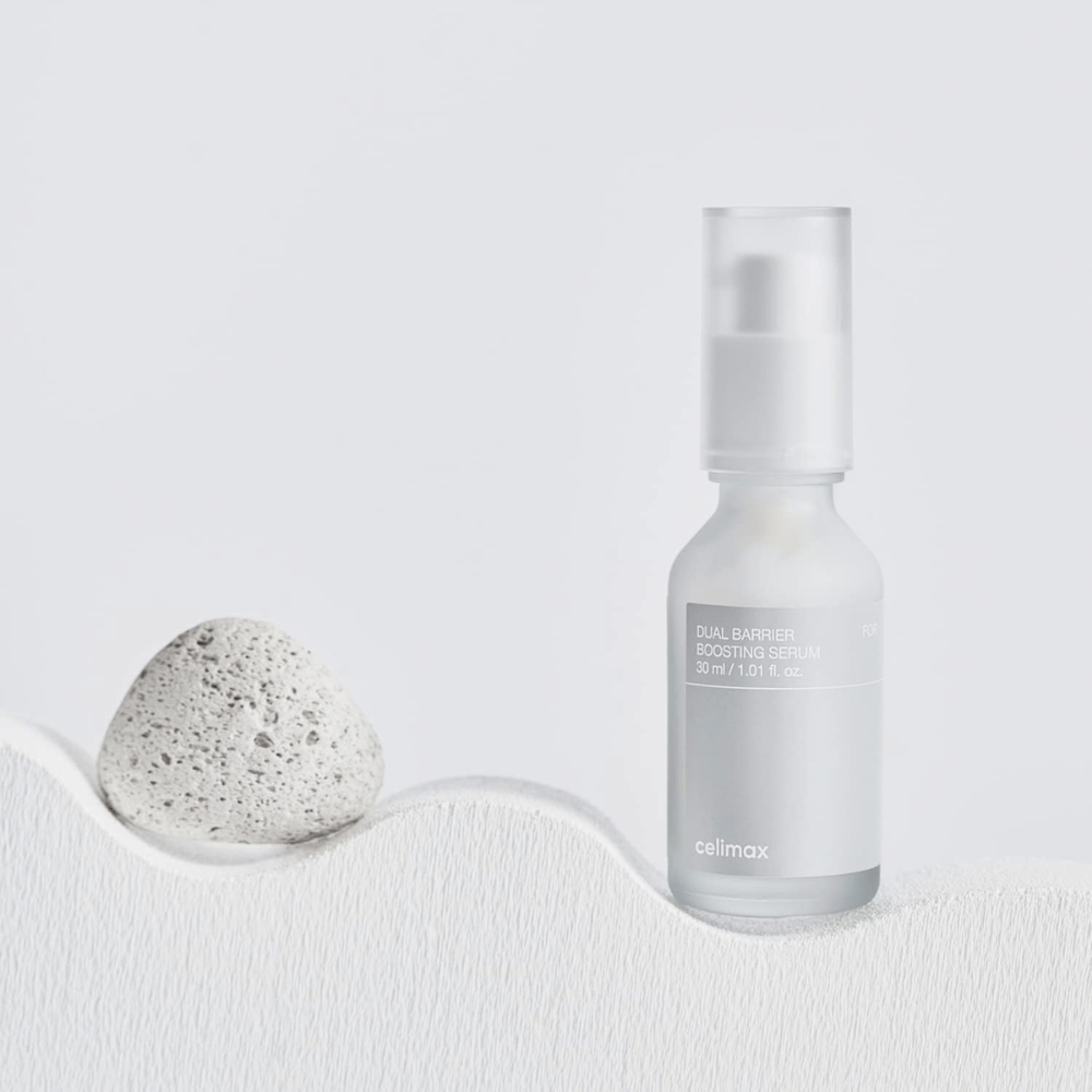 Celimax Dual Barrier Boosting Serum postavljen na tekstil s kamnom, mirna slika izdelka, ki signalizira vlaženje in okrepljeno kožno pregrado.