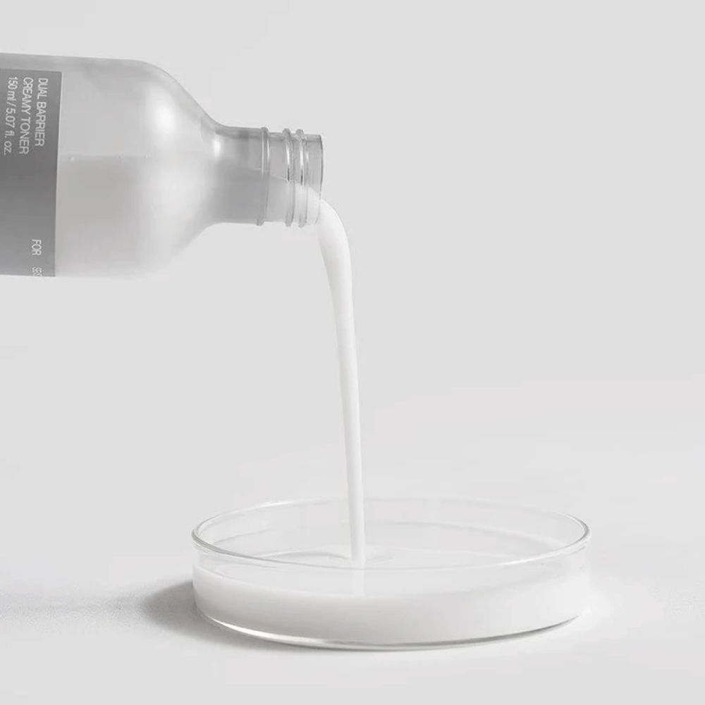 Celimax Dual Barrier Creamy Toner se previdno vlije iz steklenice v okroglo laboratorijsko posodo, pri čemer je njegova mlečna in hranljiva formula jasno v ospredju.